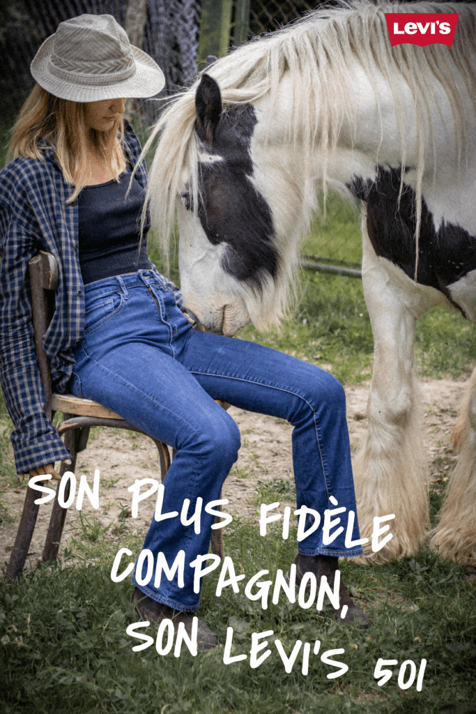 Femme blonde style cow-boy portant un jean Levi's, assise sur une chaise dans un champs proche d'un cheval qui renifle sa main. Avec le slogan "son plus fidèle compagnon son Levi's 501"